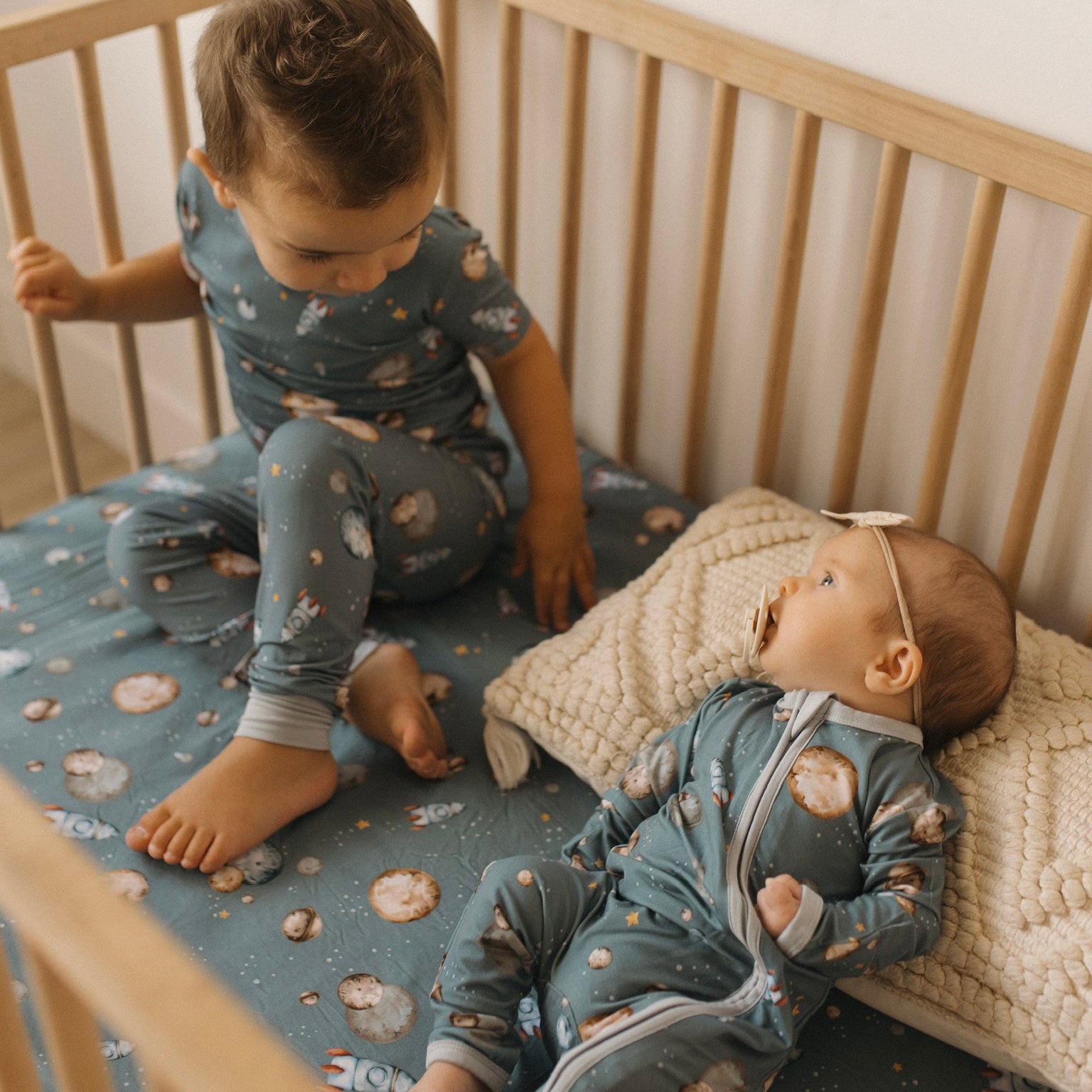 Child and baby hanging out in crib, both in space themed bamboo pajamas